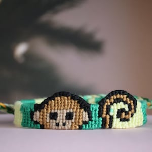 Op de afbeelding: Handgemaakte vriendschapsarmband met een apenontwerp. De armband is voornamelijk groen, geel en beige, met zwarte accenten. Het apenontwerp staat centraal, met gevlochten uiteinden om te knopen.