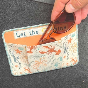 Könnte beinhalten: Eine kleine Stofftasche mit einem Meeres-Motiv und dem Text "Let the shine" ist an der Innenseite eines Autos befestigt. Ein Paar braune Sonnenbrillen liegt in der Tasche.