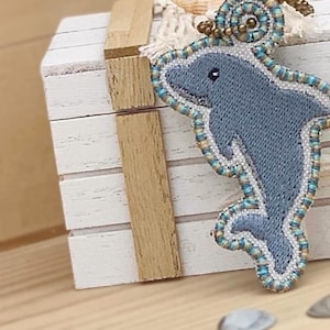 May include: A blue and white beaded dolphin charm with a brown and gold beaded accent. The charm is hanging from a white wooden box.