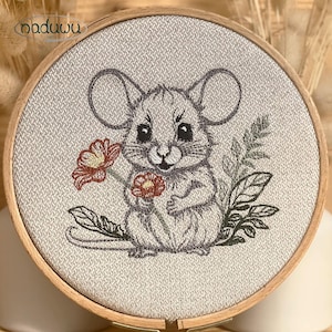 May include: Embroidered artwork of a grey mouse holding red flowers and greenery, framed in a wooden embroidery hoop. The mouse has large ears and a sweet expression. The background is a textured, light-coloured fabric.