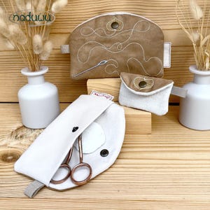 May include: A collection of handmade sewing accessories, including a brown suede-like pouch with white embroidery and a small white pouch with a button closure. A white case with scissors and a small white pouch are also visible. The items are displayed on a wooden surface.