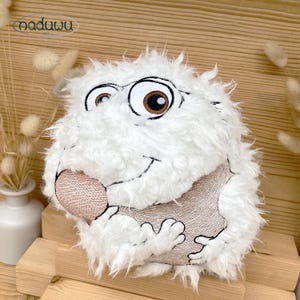 May include: A plush, white, cartoon-like creature pillow with large eyes, a brown nose, and a smiling mouth. The pillow has a textured, furry appearance and is set against a wooden background. The pillow has the word "naduuu" in the top left corner.