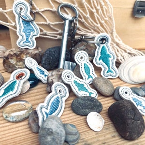 May include: A set of nine white leather keyrings with blue and green embroidered fish designs. The keyrings are scattered on a wooden surface with seashells and stones.