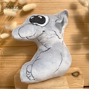 Op de afbeelding: Een lichtgrijs pluche speelgoed in de vorm van een cartoon dier, mogelijk een kat of hond. Het heeft grote zwarte ogen, een lachende mond en zwarte geborduurde details. Het speelgoed staat op een houten oppervlak.