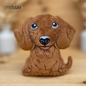 May include: A soft brown dachshund dog toy with embroidered features. The dog has large eyes, a black nose, and droopy ears. The logo "naduuu" is visible. The toy is placed on a wooden surface.