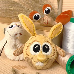 Pode incluir: Três brinquedos de pelúcia: um verme branco peludo, um coelho amarelo com olhos grandes e um caracol marrom com uma concha listrada. Os brinquedos estão em uma superfície de madeira com um carretel de fio.
