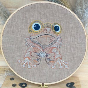 May include: Embroidered hoop art featuring a brown frog with large, round, yellow-rimmed eyes. The frog is wearing a brown, furry coat and holding a round, silver object in its hands.