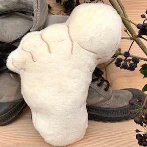 Könnte beinhalten: Ein weißes Plüschtier in Form einer Hand mit braunen Nähten. Das Spielzeug liegt auf einer Holzoberfläche neben einem Paar braunen Stiefeln.