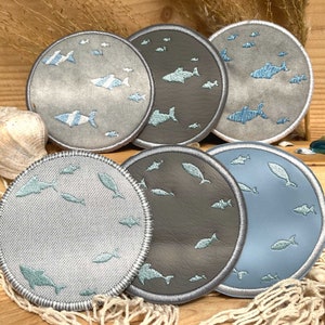 Op de afbeelding: Vijf ronde geborduurde patches met een blauw en wit visontwerp. De patches liggen op een houten oppervlak met een visnet.