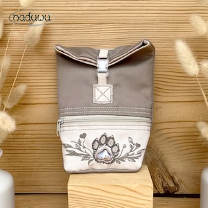 May include: A taupe and cream roll-top bag featuring a clear plastic buckle closure and a front zippered pocket. The lower cream section displays an embroidered design of a paw print surrounded by leafy vines. The brand name naduwi is visible.
