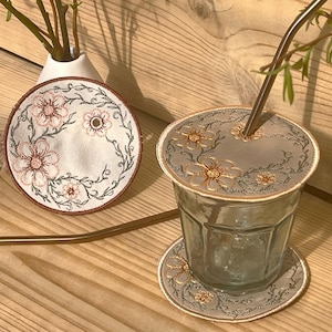 May include: Two embroidered coasters featuring a floral design. One coaster is white with pink and brown embroidery, the other is beige with gold embroidery. A glass with a straw is sitting on the beige coaster.
