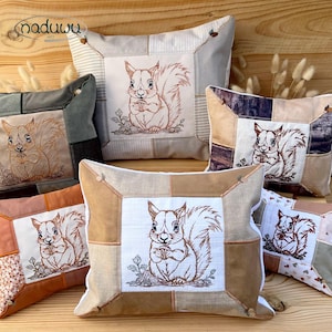 May include: Several decorative cushions with embroidered squirrel motifs. The pillows have a patchwork design in neutral tones like beige, brown, and grey. The squirrel embroidery is in brown and cream shades. The cushions are arranged on a wooden surface.
