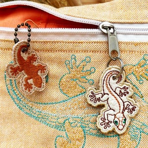 May include: A beige fabric bag with a zipper closure and two embroidered gecko charms. One charm is brown and the other is white with green eyes.