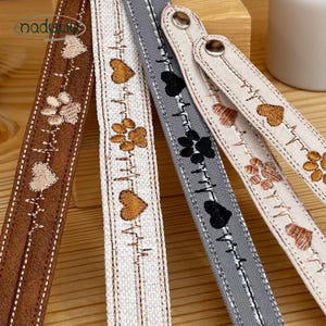 May include: Four fabric wristbands featuring embroidered heart and paw print designs connected by a heartbeat line. The bands come in brown, white, and gray, each secured with a metallic snap button. The word naduwy is printed in the top left corner.