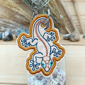 May include: A beige embroidered gecko keychain with a brown tail, blue eyes, and orange accents. The keychain is attached to a silver key ring.