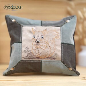 May include: Square cushion with a patchwork design in shades of grey and teal. The centre features an embroidered squirrel holding a nut. The cushion has the text "naduuu ART" in the upper left corner.