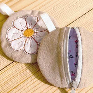 May include: Two beige, round pouches. One features a large embroidered daisy with white petals and a yellow center. The other pouch is open, revealing a floral patterned interior. Both have a white zipper and a white fabric loop.