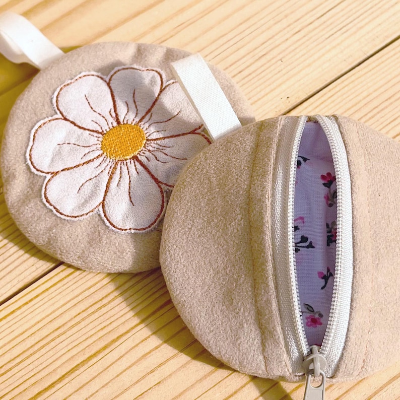 May include: Two beige, round pouches. One features a large embroidered daisy with white petals and a yellow center. The other pouch is open, revealing a floral patterned interior. Both have a white zipper and a white fabric loop.