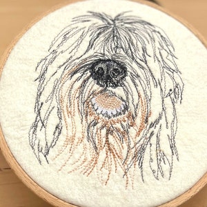 Op de afbeelding: Borduurwerk van een hondenkop op een witte stoffen achtergrond. De hond heeft zwart en bruin haar en een witte neus.