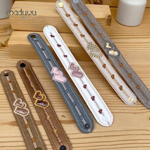 Puede incluir: Una colección de pulseras decorativas en varios colores, incluyendo marrón, gris y blanco. Cada pulsera presenta diseños de corazones bordados y pequeños detalles de corazones. Las pulseras se exhiben sobre una superficie de madera.