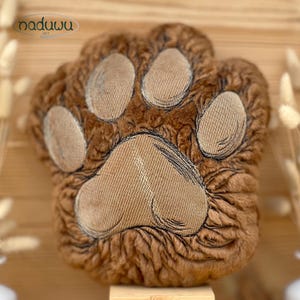 May include: A brown plush cushion shaped like an animal paw, featuring a textured faux fur surface and four smaller oval-shaped pads above one large central pad, all in a lighter tan colour with embroidered outlines and the text naduwu art.