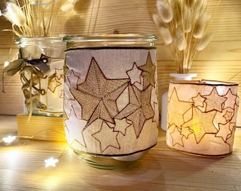 Puede incluir: Frascos de vidrio decorados con patrones de estrellas doradas. Los frascos están envueltos con una tela color crema bordada con estrellas doradas. Los frascos están iluminados con una luz suave y cálida, creando un ambiente acogedor. Flores secas en jarrones en el fondo.