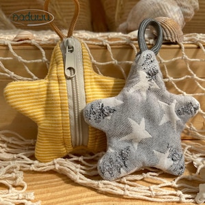 May include: Two star-shaped fabric pouches with zipper closures and hanging loops. One pouch is yellow with white horizontal stripes, and the other is gray with a white star pattern. The yellow pouch features a beige zipper and a tan loop.