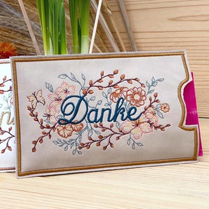 Könnte beinhalten: Eine beige Karte mit einem gestickten Blumendesign und dem Wort "Danke" in türkisblauer Schrift. Das Blumenarrangement umfasst rosa und pfirsichfarbene Blüten, braune Zweige und türkisfarbene Blätter. Die Karte hat einen goldenen Rand.