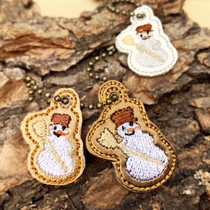 May include: Three embroidered snowman ornaments with brown felt hats and white faces. The ornaments are attached to gold chains and are displayed on a piece of tree bark.