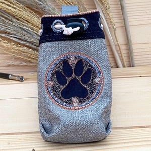 Könnte beinhalten: Eine graue Kordeltasche mit einem gestickten schwarzen Pfotenabdruck auf der Vorderseite. Die Tasche hat einen schwarzen und weißen Kordelverschluss und einen schwarzen Riemen.