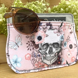 Könnte beinhalten: Ein rosa Stoff-Sonnenbrillenetui mit floralem Muster und Totenkopf. Das Etui hat einen grauen Rand und ein kleines Fach zum Verstauen der Sonnenbrille.