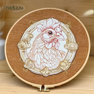 Könnte beinhalten: Gesticktes Kunstwerk eines Hahns in einem Holzreifen. Der Hahn ist in rotem und weißem Faden dargestellt, vor einem braunen Stoffhintergrund. Das Kunstwerk ist mit einem goldfarbenen Zierrahmen versehen. Der Reifen ist mit einem Messingverschluss befestigt.