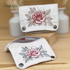 Könnte beinhalten: Zwei cremefarbene Leinentaschen mit einer gestickten roten Rose und grünen Blättern. Jede Tasche hat eine Klappe, die mit zwei metallischen Druckknöpfen verschlossen wird. Die vordere Tasche ist geöffnet und zeigt ein helles Innenfutter.