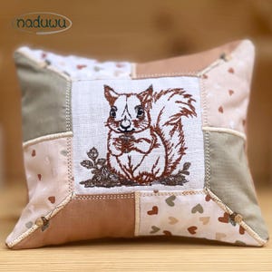 May include: A square cushion with a patchwork design. The central panel features an embroidered brown squirrel holding a nut. The cushion has a beige, brown, and green colour scheme, with heart-shaped patterns on some sections. The brand name "naduwu" is visible.