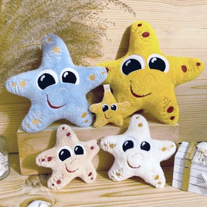 Pode incluir: Quatro brinquedos de pelúcia em forma de estrela do mar de diferentes tamanhos e cores. A maior estrela do mar é amarela com detalhes vermelhos, a próxima maior é azul com detalhes brancos, a próxima é bege com detalhes vermelhos e a menor é branca com detalhes dourados. Todas as estrelas do mar têm olhos e sorrisos bordados.