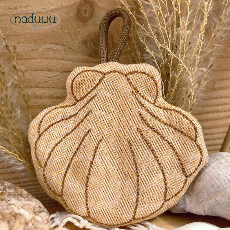 Pode incluir: Uma bolsa de tecido bege em forma de concha com uma trama texturizada e detalhes costurados em castanho ao longo das nervuras. A bolsa possui uma al&ccedil;a de cord&atilde;o castanho no topo para pendurar. A marca naduwu est&aacute; impressa no canto superior.