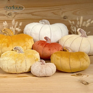 May include: A collection of six plush pumpkin-shaped cushions in various sizes and colours, including white, cream, orange, and yellow. Each soft fabric pumpkin features stitched vertical segments and a brown stem. The brand name naduwu is visible.