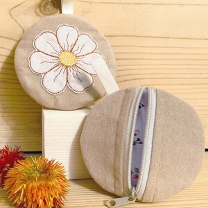 May include: Two beige round coin purses. One features a white daisy embroidery with a yellow center. The other is open, revealing a floral patterned interior. Both have a white zipper and a small loop.