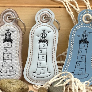 May include: Three keychains with a lighthouse design. The keychains are made of fabric and leather. One keychain is white with brown stitching, one is light blue with white stitching, and one is beige with brown stitching.