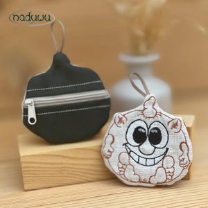 May include: Two small, handmade coin purses. One is black with a silver zip and a loop for hanging. The other is white with a smiling cartoon face embroidered in black and brown. The purses are on a wooden block.