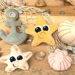 May include: A set of five plush toys shaped like a blue anchor, two starfish, and two seashells. The starfish are yellow and cream coloured, and the seashells are white and pink.