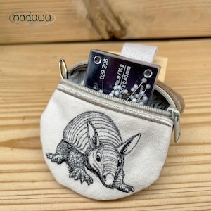 May include: A small, beige pouch with a zipper, featuring an embroidered armadillo design. The pouch contains a package of sewing needles. The text "029 208" and "No. 9 / 10g 30 x 0,60 mm" are visible on the needle package.