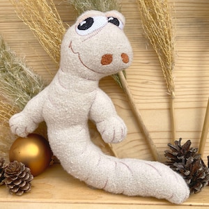 以下が含まれることがあります： 笑顔と大きな目をした白いぬいぐるみワーム。ワームは松ぼっくりと金色のボールのある木製の表面に横たわっています。