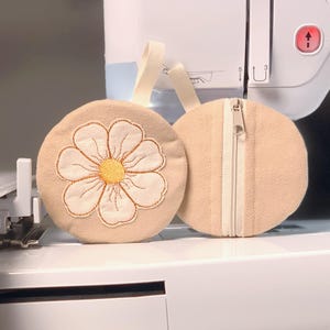 May include: Two beige, round pouches with cream-colored daisy embroidery and a zipper. One pouch features a daisy design with a yellow center and cream petals. The other pouch has a cream-colored zipper.
