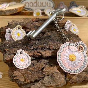 Könnte beinhalten: Ein silberner Schlüsselanhänger mit einem Schlüssel und drei gestickten Gänseblümchen-Anhängern. Die Anhänger sind rosa, weiß und gelb. Die Anhänger sind an einer silbernen Kette und einem silbernen Schlüssel befestigt.