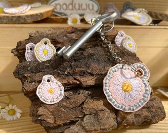 May include: A silver key chain with a key and three embroidered daisy charms. The charms are pink, white, and yellow. The charms are attached to a silver chain and a silver key.
