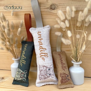 以下が含まれることがあります： 刺繍のディテールと吊り下げループが付いた、アルマジロをテーマにした長方形の布製サシェ3つ。サシェは緑、オフホワイト、タンで、それぞれに「armadillo」の文字が同系色で刺繍されています。サシェはドライフラワーと一緒に展示されています。