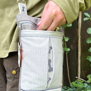 May include: A gray and white striped crossbody bag with a zipper closure and a small pocket on the front. The bag has a gray strap and a silver zipper pull.
