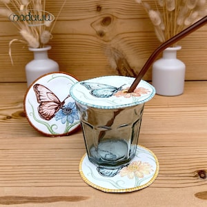 Könnte beinhalten: Ein Glas mit einem Deckel und Untersetzer mit Schmetterlingsmotiven, bestickt mit Schmetterlingen und Blumen in Blau, Orange und Braun. Ein brauner Strohhalm steckt im Glas. Das Wort "naduwa" ist oben links zu sehen.