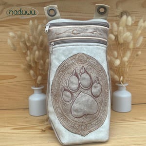 Può includere: Borsa grigio chiaro con un grande design di impronta di zampa ricamata. La borsa ha due scomparti con cerniera e due cinturini beige con occhielli metallici. È decorata con un bordo ricamato marrone. Il marchio "naduuu" è visibile.